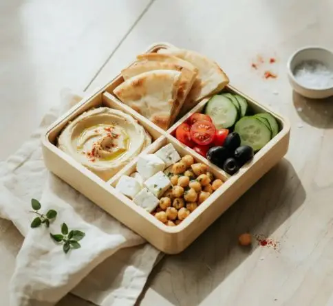 Greek mezze bento with hummus, pita, cucumber, tomatoes, feta, chickpeas, and olives.