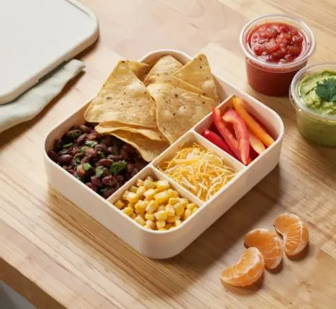 DIY nacho bento with tortilla chips, black beans, cheese, corn, peppers, salsa, guacamole, and mandarins.