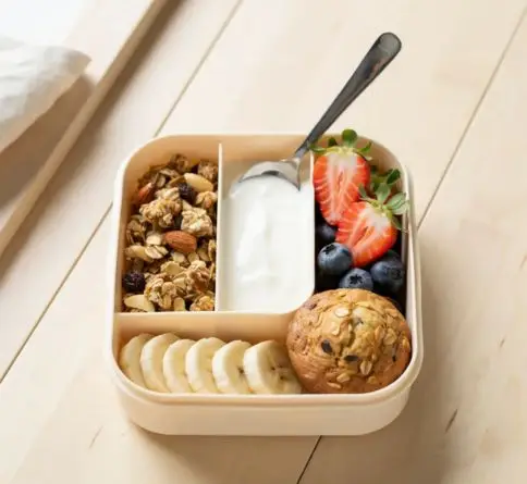 Yogurt granola fruit bento with Greek yogurt, granola, berries, banana, and a mini muffin.